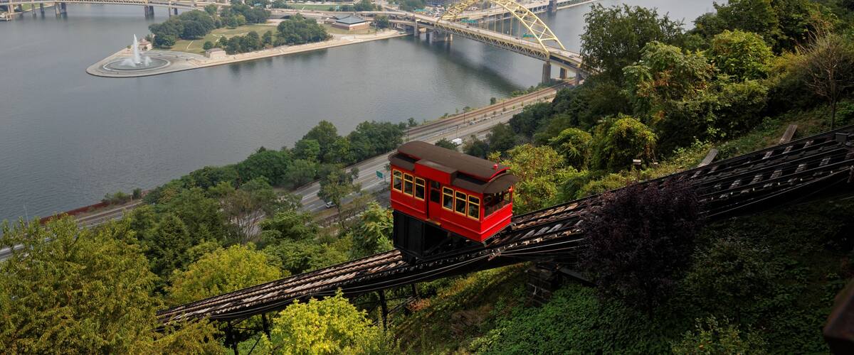 A red cable car on tracks, a river, a city skyline with tall buildings, and a bridge.