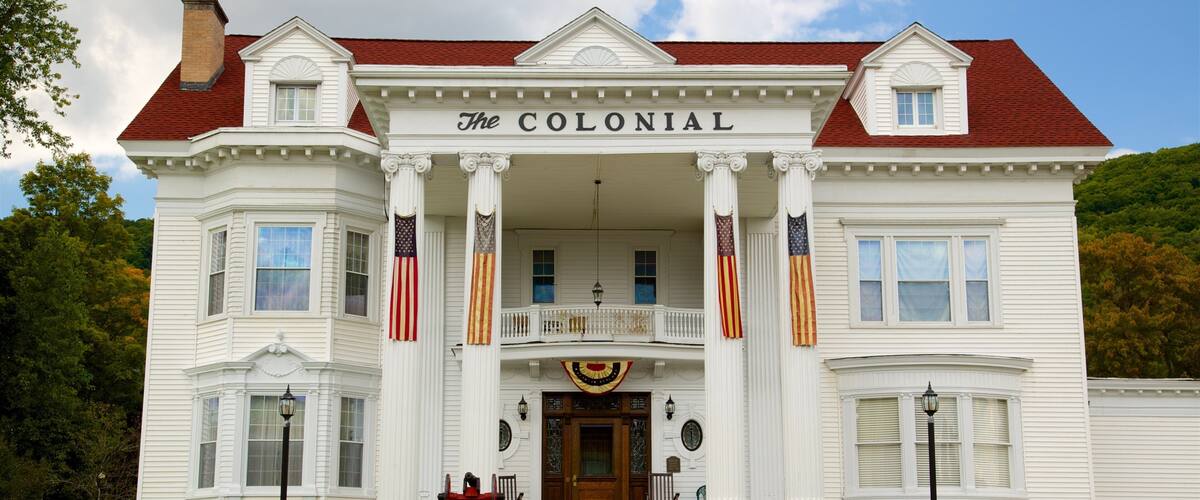 A large white colonial-style house with a red roof, named "The Colonial," featuring a front porch with columns, American flags, and a central entrance.