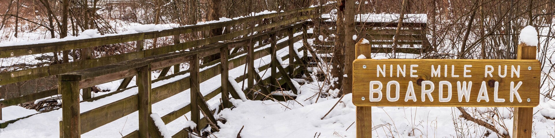 A wooden sign engraved with Nine Mile Run Boardwalk, a stream and walkway in Frick Park, an urban park in Pittsburgh, Pennsylvania, USA as seen on a sunny winter day