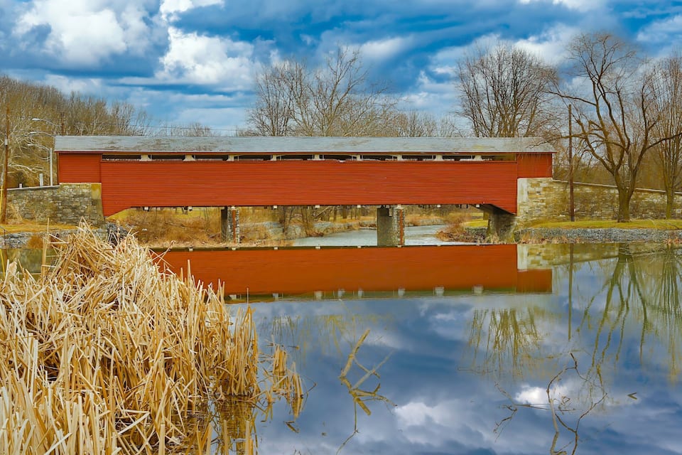 A red covered bridge over a calm pond with reflections, surrounded by trees and dry reeds.
