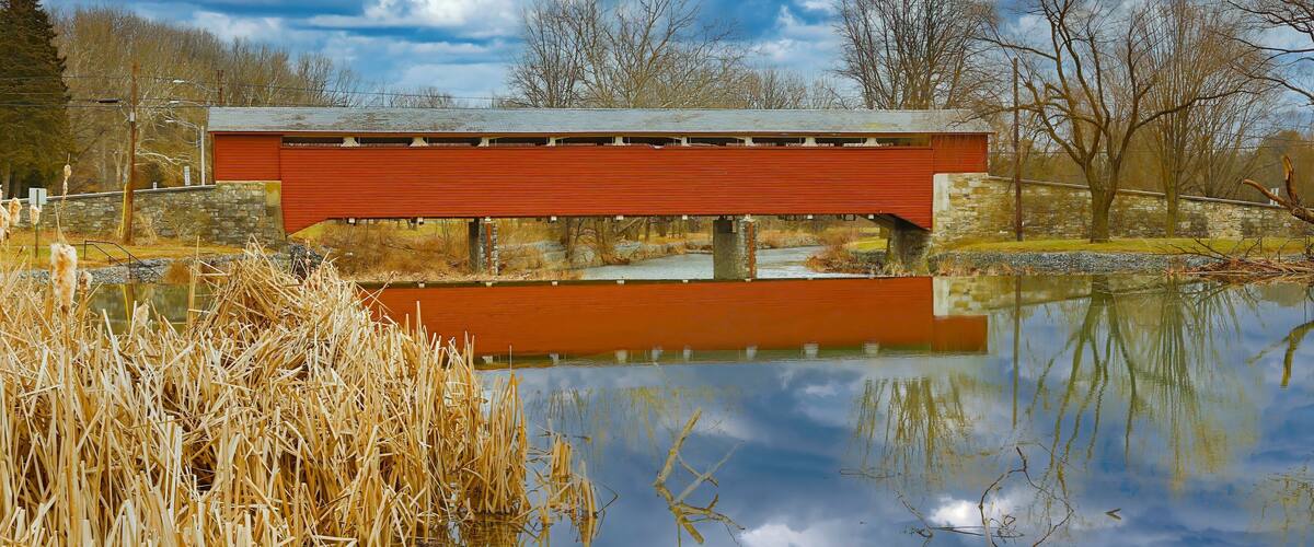 A red covered bridge over a calm pond with reflections, surrounded by trees and dry reeds.