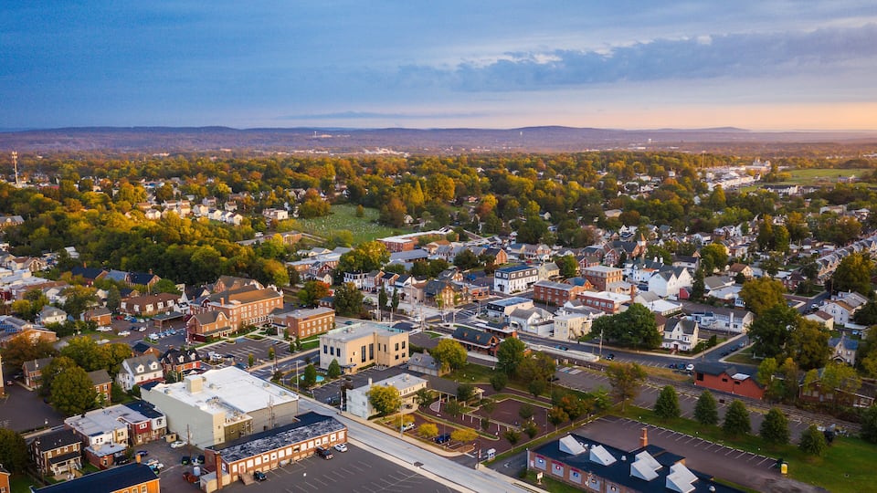 An aerial view of a town with a mix of residential and commercial buildings, surrounded by green areas and hills in the background.