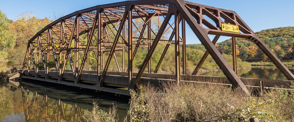 A rusted metal bridge over a calm river with autumn foliage in the background.