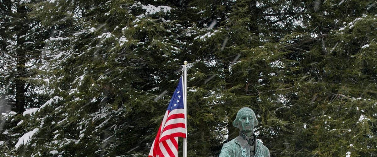 A statue of a historical figure with an American flag in a snowy park with benches and trees.
