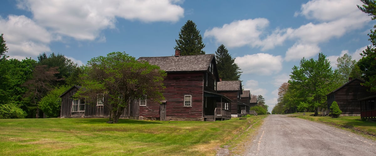 A rural road flanked by wooden houses and green fields under a partly cloudy sky.