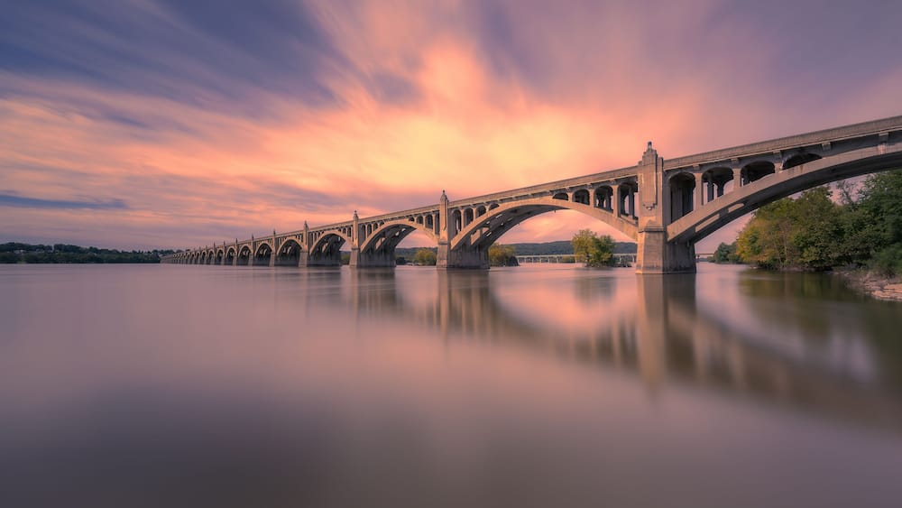 Un long pont à arches multiples enjambant une rivière paisible, entouré d’arbres et illuminé par un ciel au coucher du soleil.