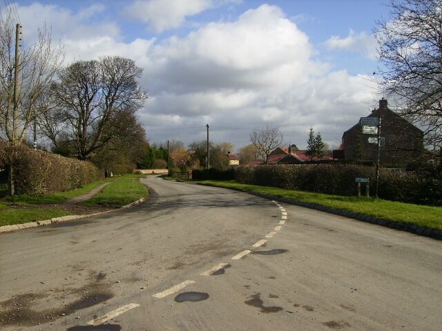 A rural road with a curve, surrounded by hedges and trees, leading to a building with a red roof.