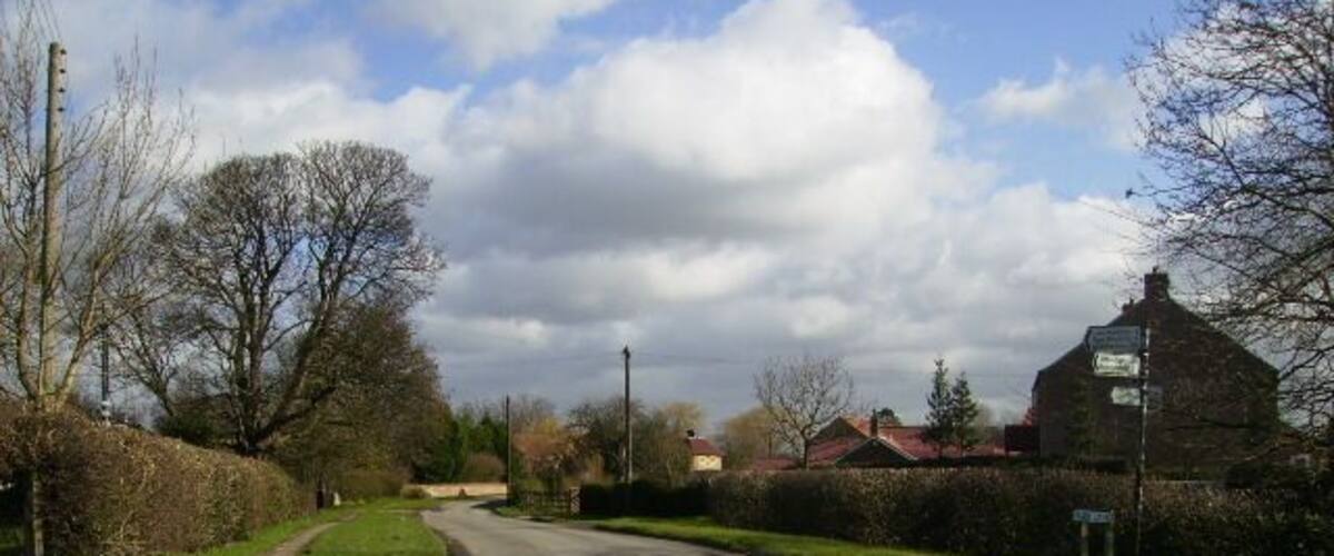 A rural road with a curve, surrounded by hedges and trees, leading to a building with a red roof.
