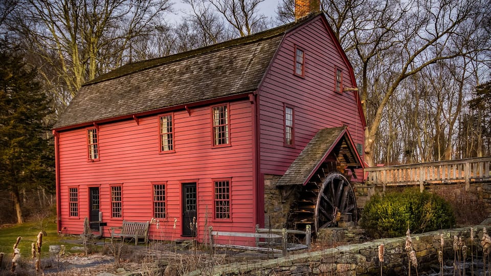 A red wooden mill with a water wheel, surrounded by trees and a stone wall.