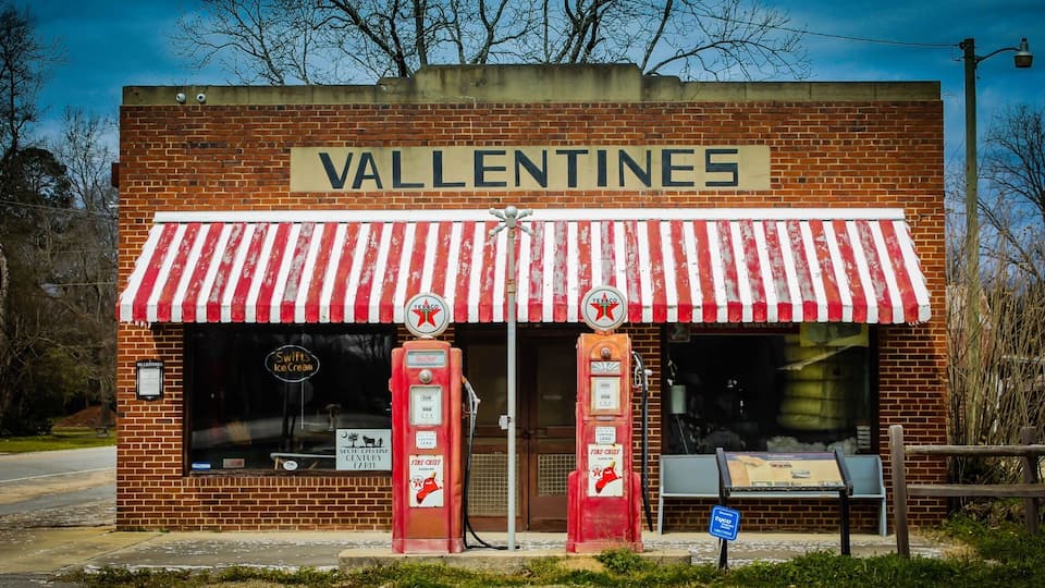 A vintage ice cream parlor named 'Valentines' with a red and white striped awning, two vintage gas pumps, and a small signboard.