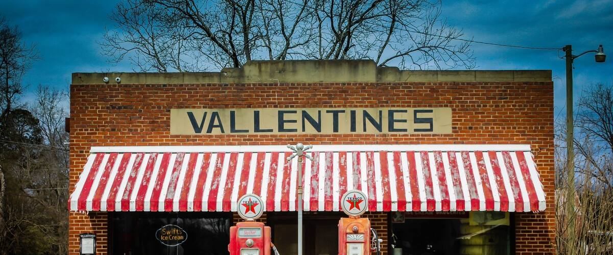 A vintage ice cream parlor named 'Valentines' with a red and white striped awning, two vintage gas pumps, and a small signboard.