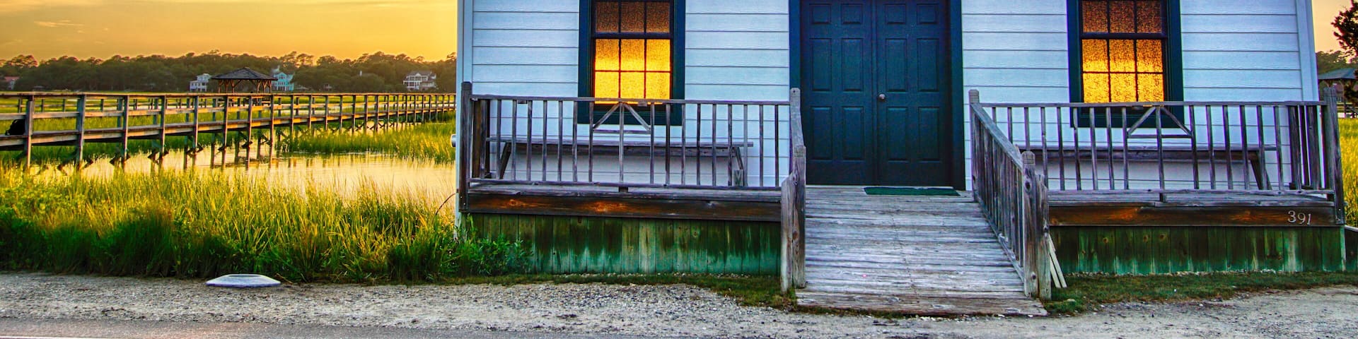 small white wooden chapel on the water on the coast during a colorful summer sunset with illuminated golden windows and a setting sun under a blue sky with soft clouds