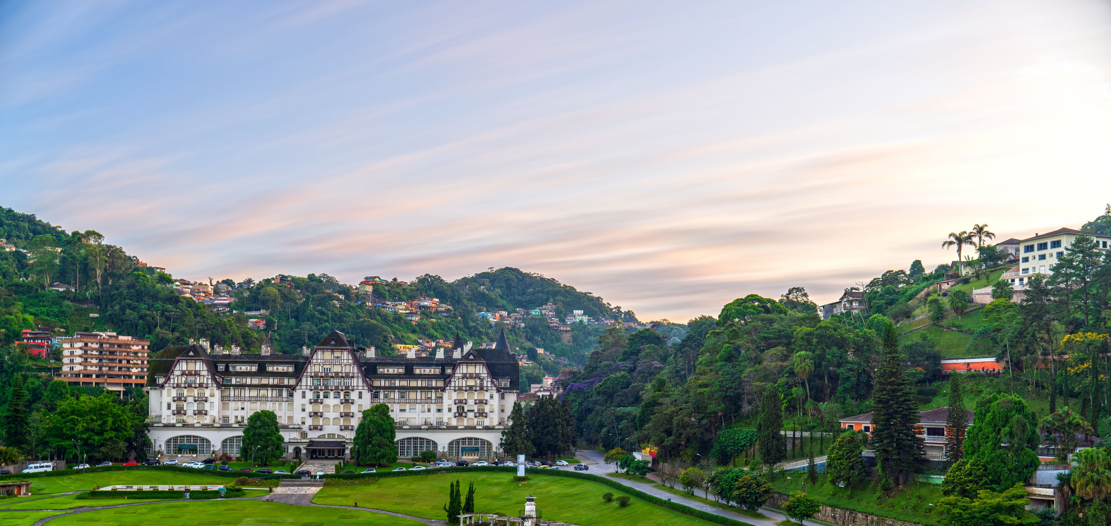 A large, historic building with a garden in front, surrounded by lush greenery and hills.