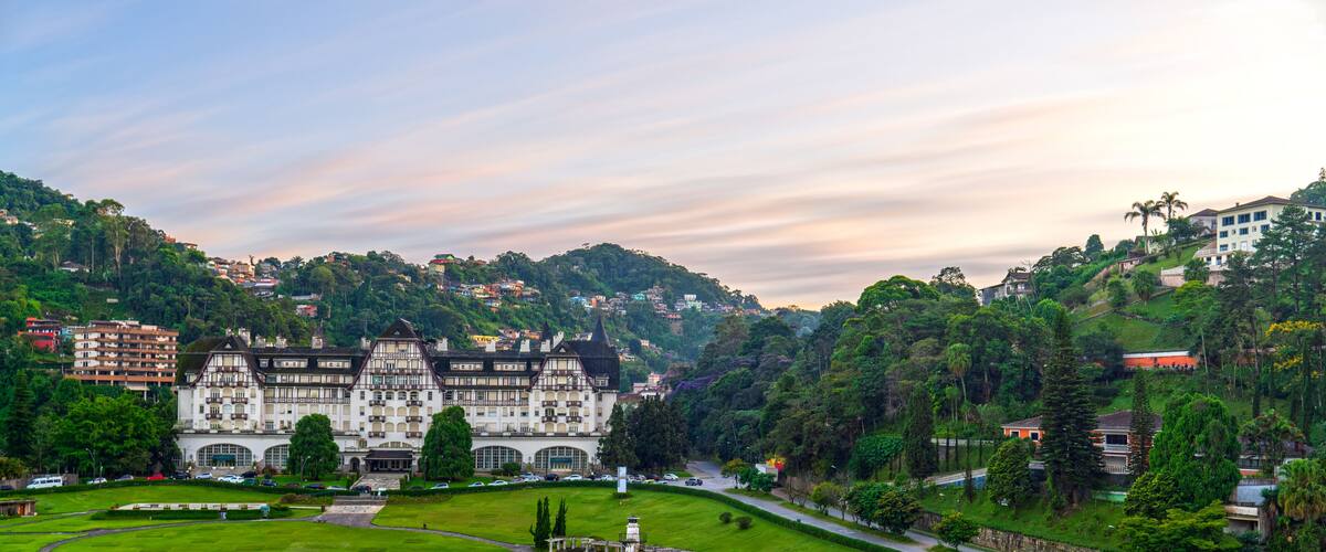 A large, historic building with a garden in front, surrounded by lush greenery and hills.