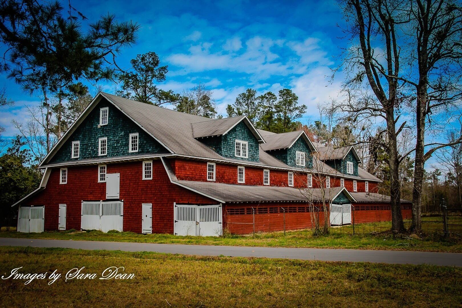 A large, two-story red barn with white trim and a green roof, surrounded by trees and a clear sky.
