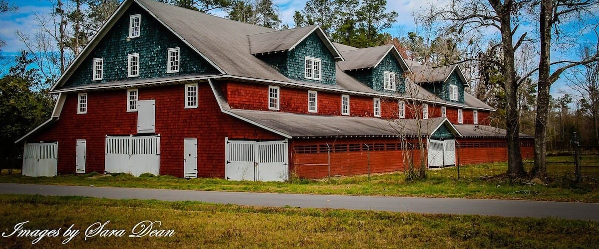 A large, two-story red barn with white trim and a green roof, surrounded by trees and a clear sky.