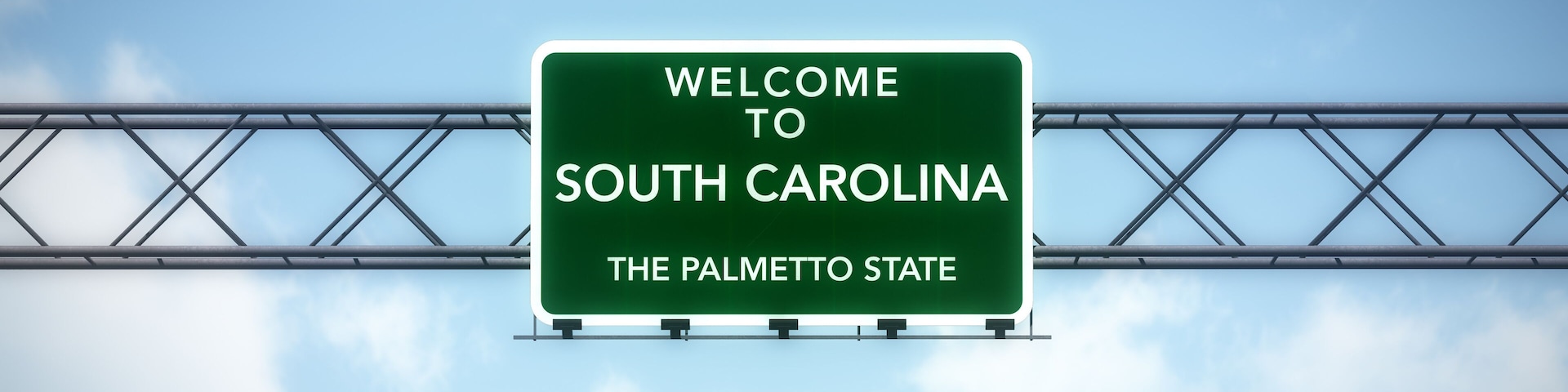 A highway sign welcoming visitors to South Carolina, The Palmetto State, against a backdrop of a clear sky and a metal bridge structure.