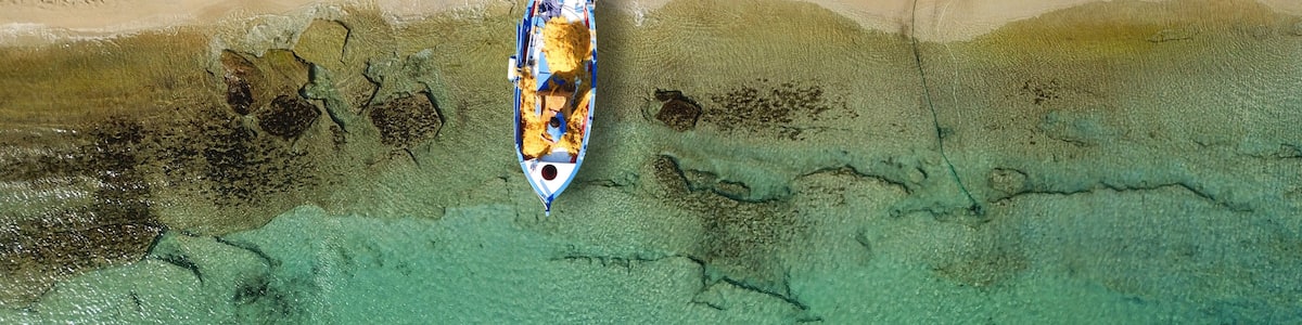 Vue aérienne d’une plage avec des transats et des parasols, un bateau sur l’eau et une eau claire et peu profonde.