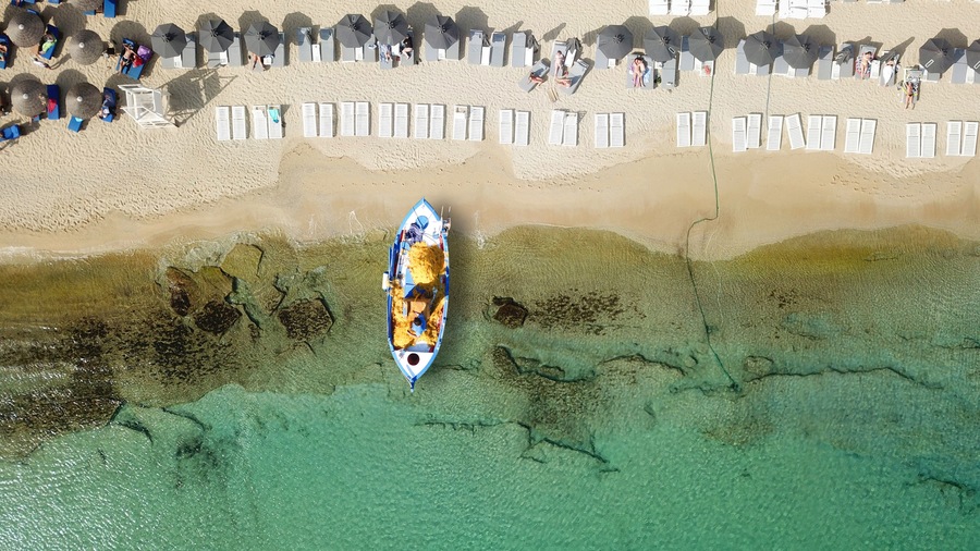 Vista aérea de uma praia com espreguiçadeiras e guarda-sóis, um barco na água e águas rasas e cristalinas.