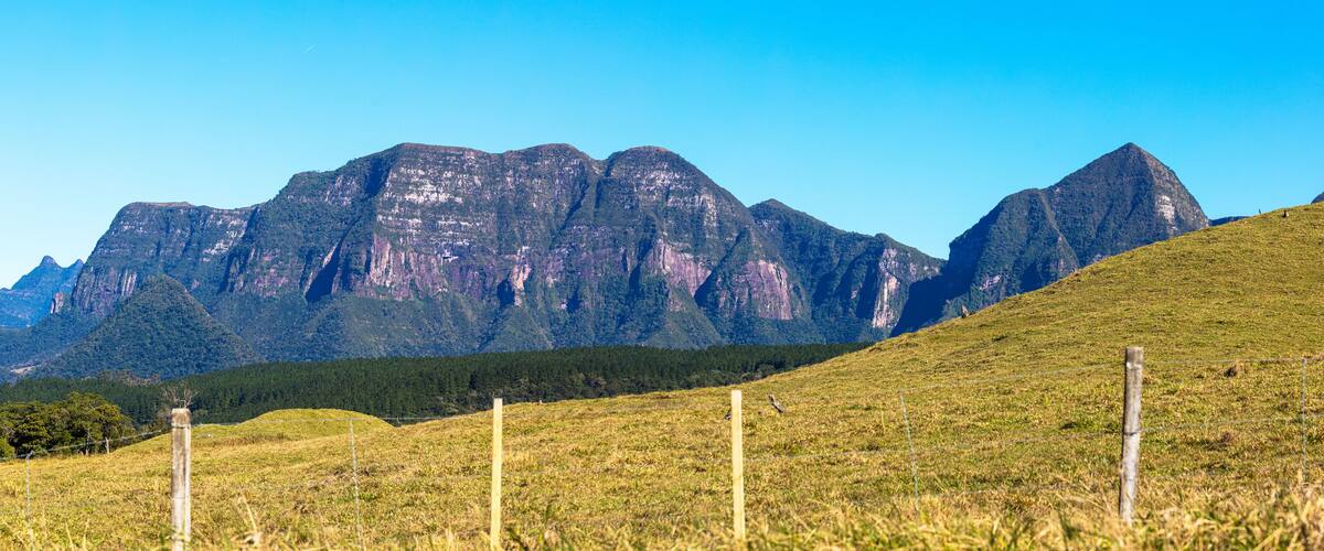 A grassy field with a wooden fence in the foreground and a mountain range in the background.
