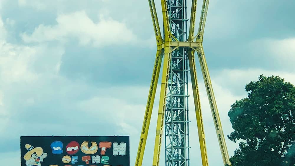 Une grande attraction foraine jaune, surmontée d’une structure colorée, entourée de verdure et se dressant sous un ciel nuageux.