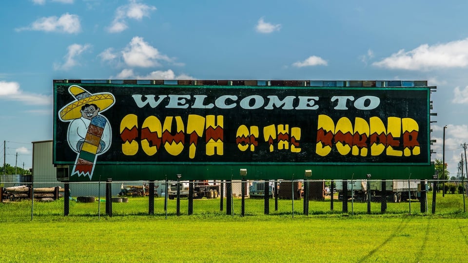 A large billboard with a cartoon character and the text "WELCOME TO SOUTH OF THE BORDER".