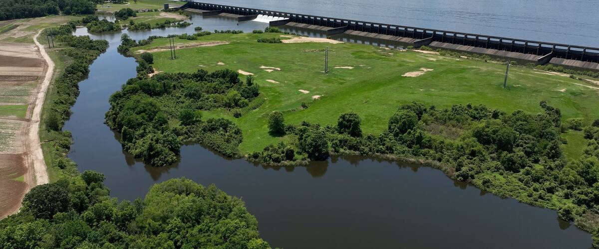 A river with a dam and a bridge, surrounded by greenery and fields.