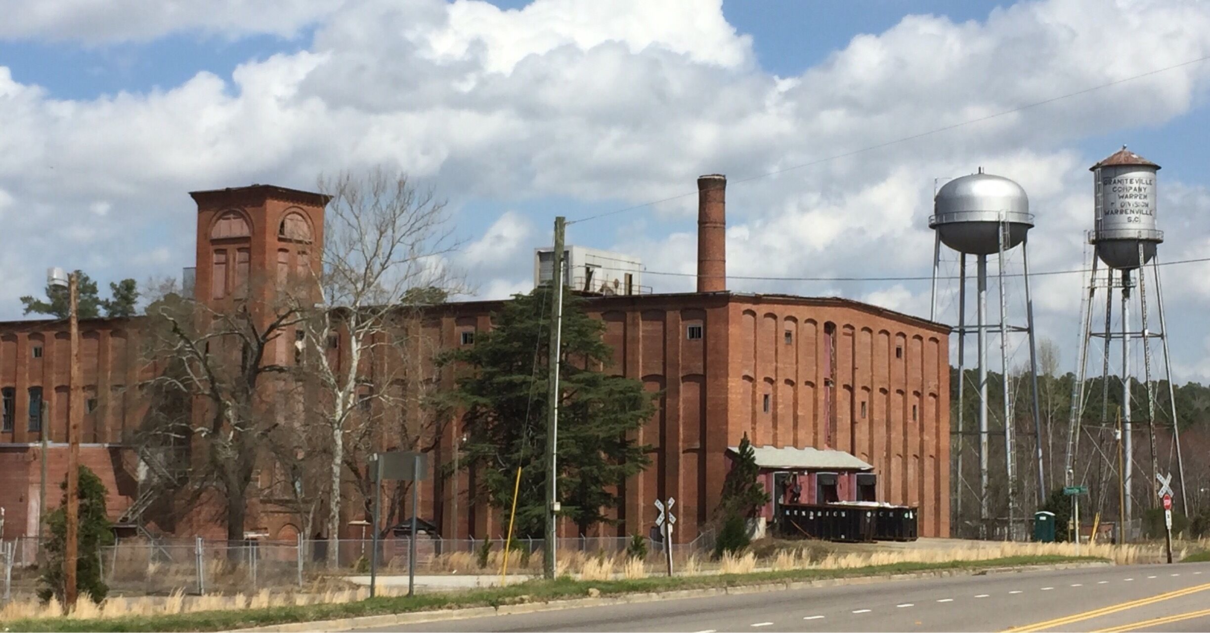 A large brick building with a tower, water towers, and a fence along a road.