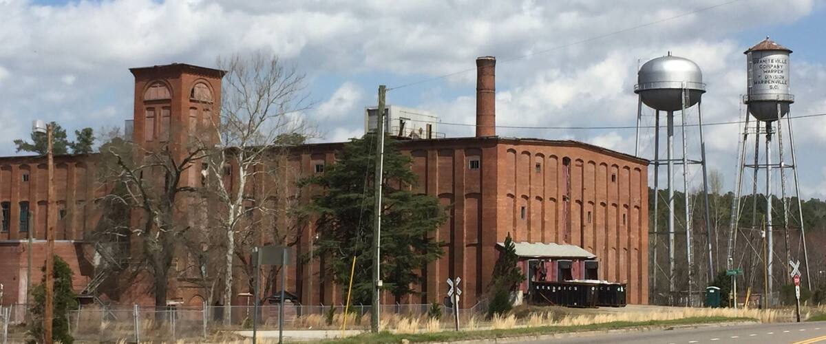 A large brick building with a tower, water towers, and a fence along a road.