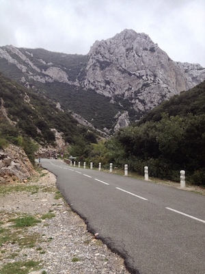 A winding mountain road with guardrails, surrounded by rocky terrain and dense vegetation.