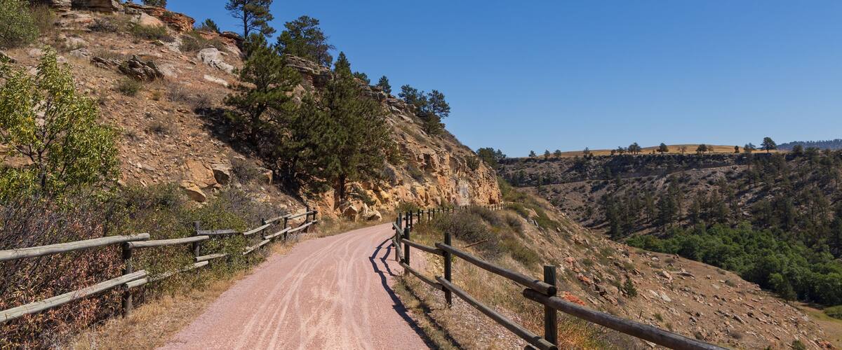 A dirt road winding through a rocky hillside with sparse vegetation and wooden guardrails.