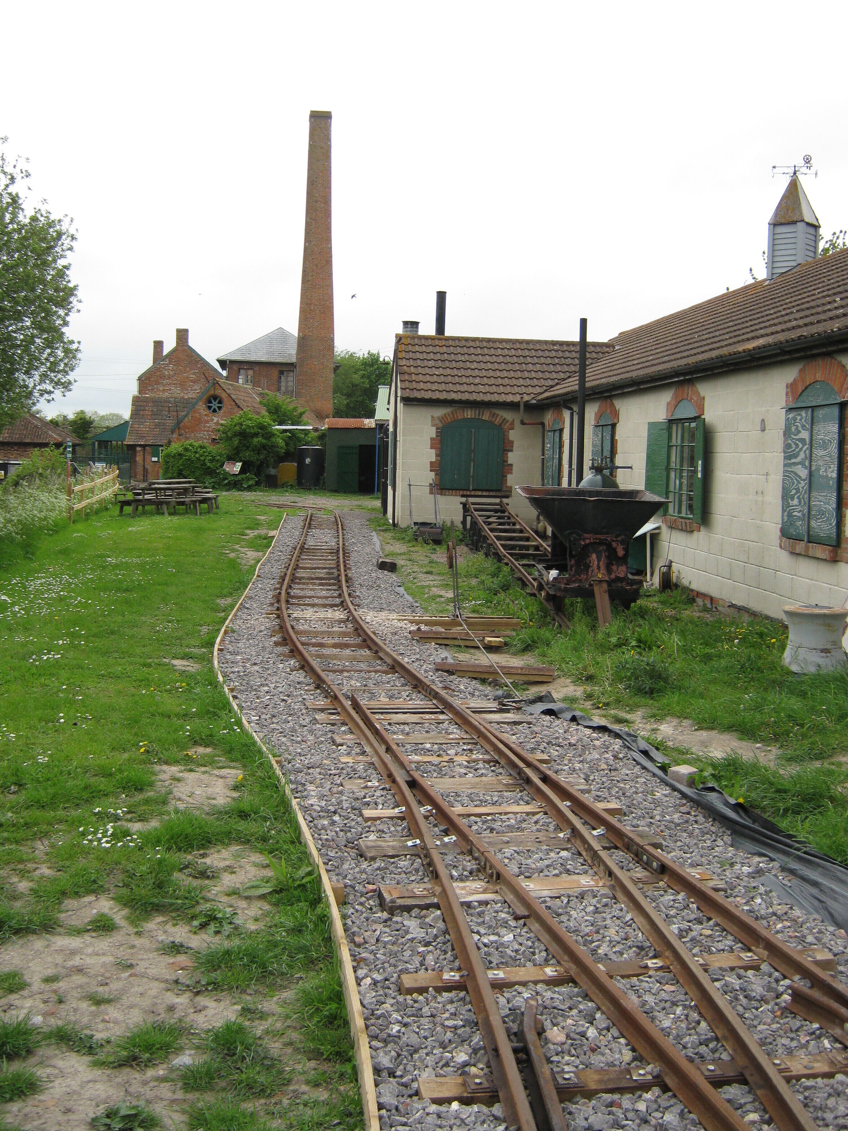 A railway track curving through a grassy area with old buildings and a tall chimney in the background.