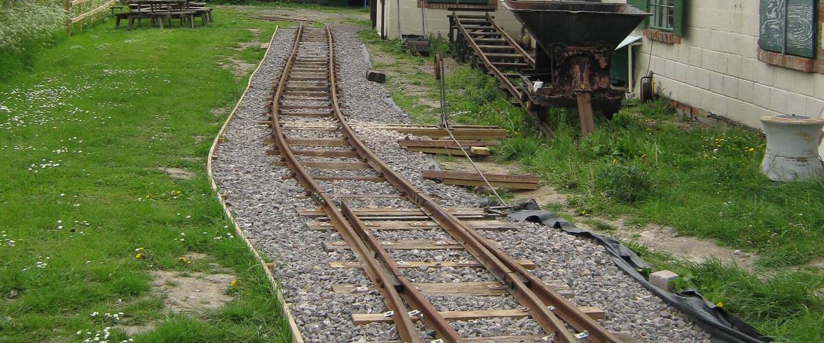 A railway track curving through a grassy area with old buildings and a tall chimney in the background.