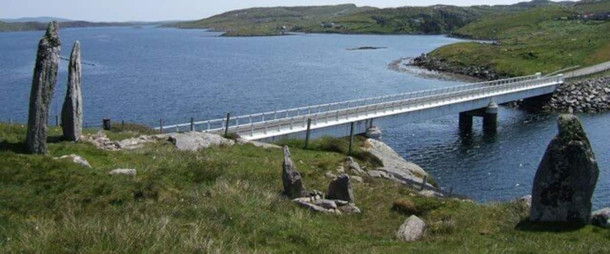 A bridge over a body of water, surrounded by grassy hills and standing stones.