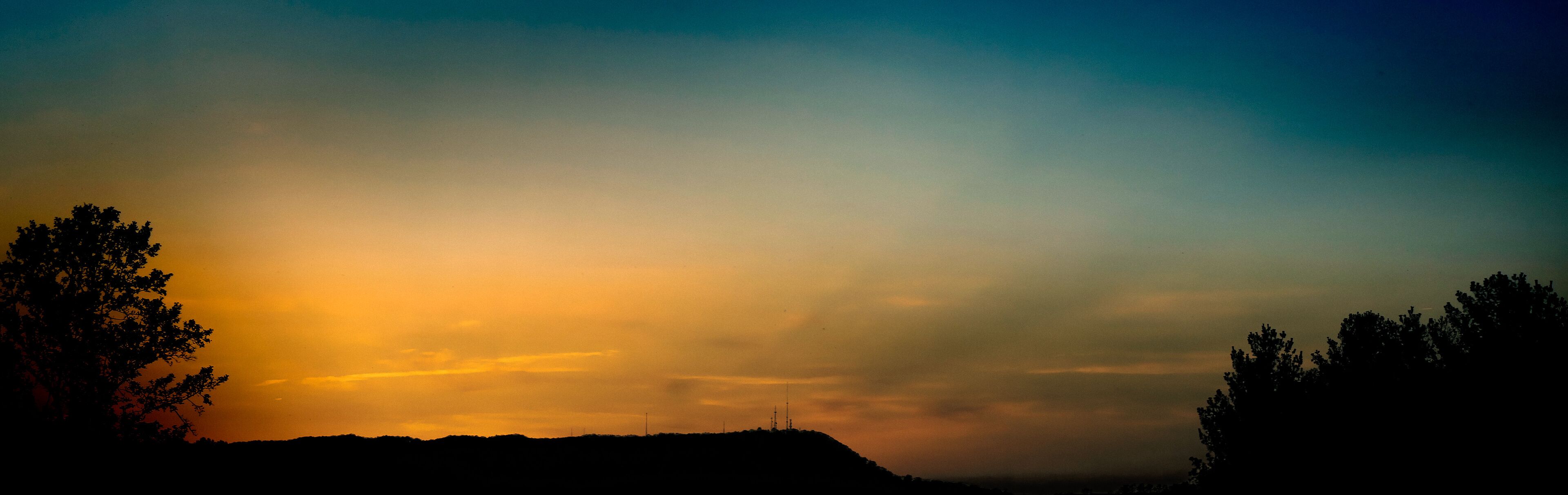 A sunset over a hilly landscape with silhouettes of trees and a distant tower.