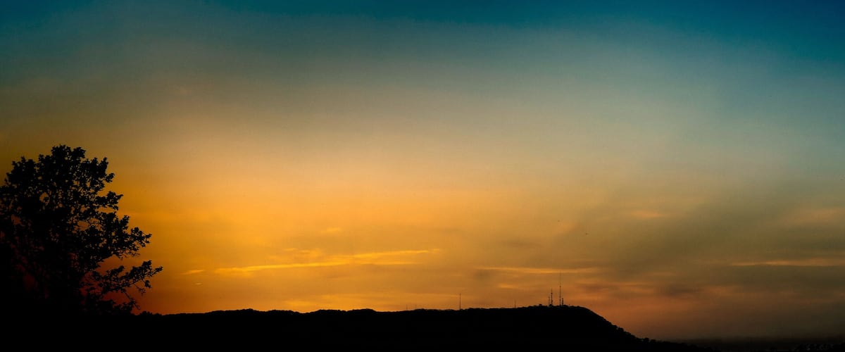 A sunset over a hilly landscape with silhouettes of trees and a distant tower.