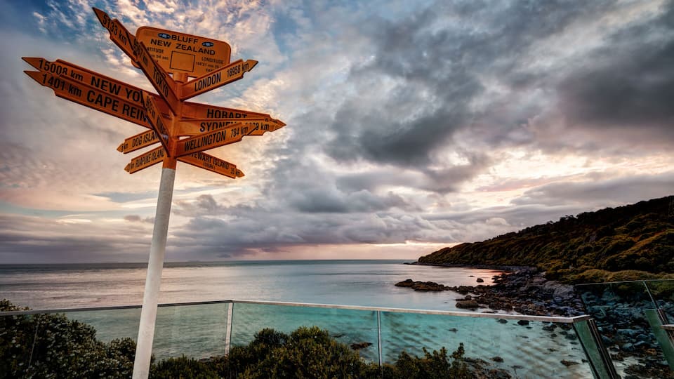 A directional signpost with destinations and distances, overlooking a coastal landscape with a view of the sea and a cloudy sky.