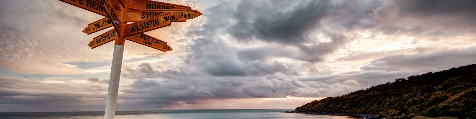 Un cartel indicador muestra destinos y distancias, con vistas a un paisaje costero y al mar bajo un cielo nublado.