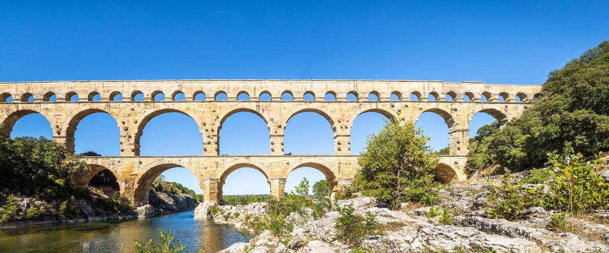 A stone aqueduct with multiple arches spanning a river, surrounded by rocky terrain and greenery.