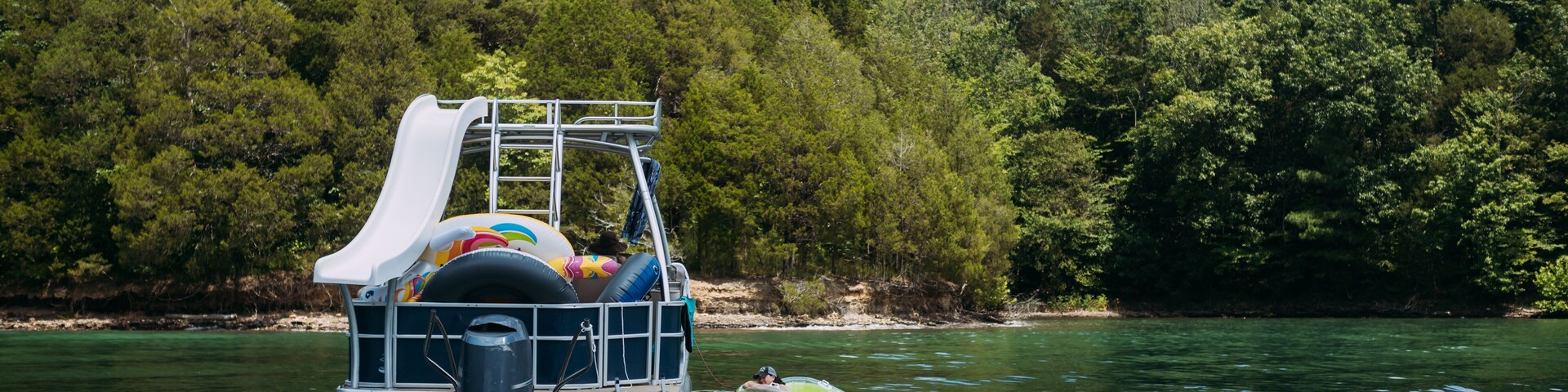 A watercraft with a slide and inflatable rings, floating on a calm river surrounded by dense forest.
