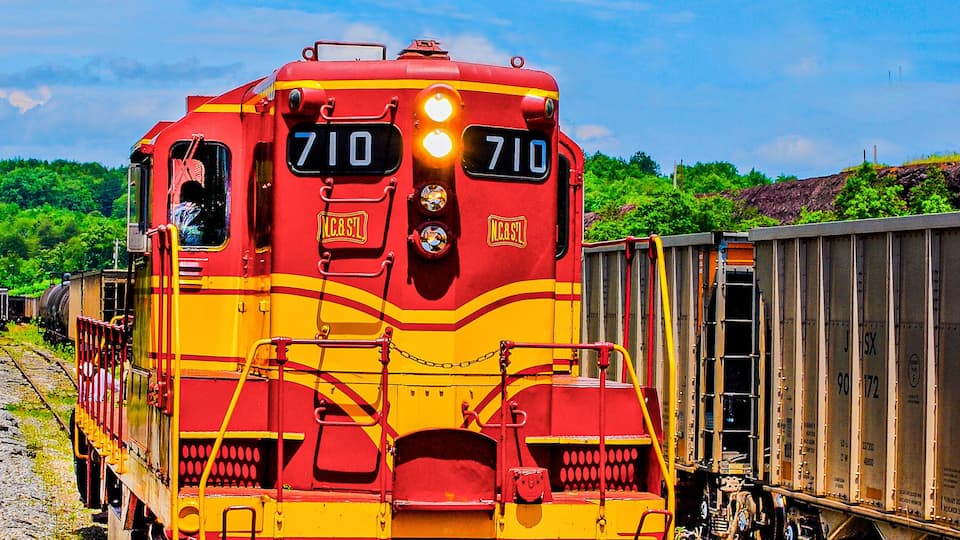 A red and yellow train with the number 710 on the front, traveling on tracks with gravel and greenery in the background.