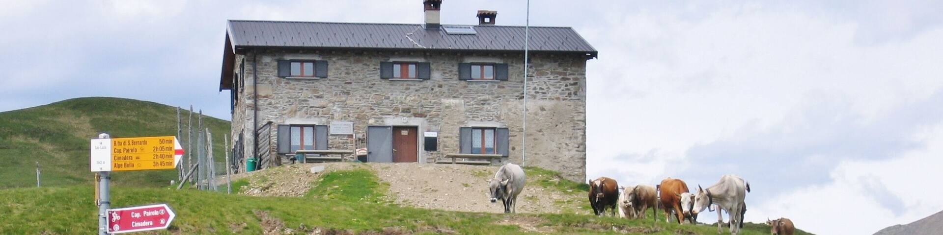 A stone building on a hillside with a signpost and grazing cows.