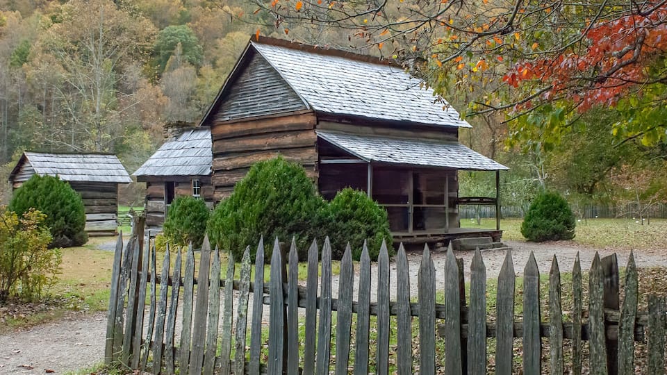 A rustic log cabin with a porch, surrounded by a wooden fence and trees with autumn leaves.