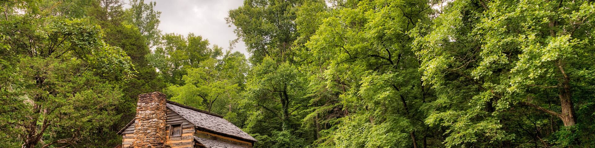 An old log and stone cabin in the green woods with an old wooden fence in the foreground and a grey sky