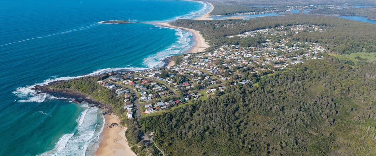 An aerial view of a coastal town with a beach, dense forest, and a river.