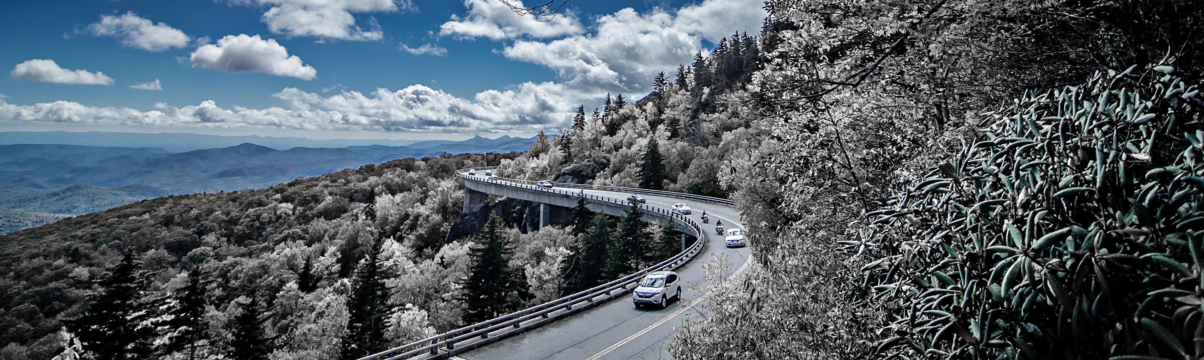 A winding mountain road with vehicles, surrounded by dense forest and distant mountains under a partly cloudy sky.