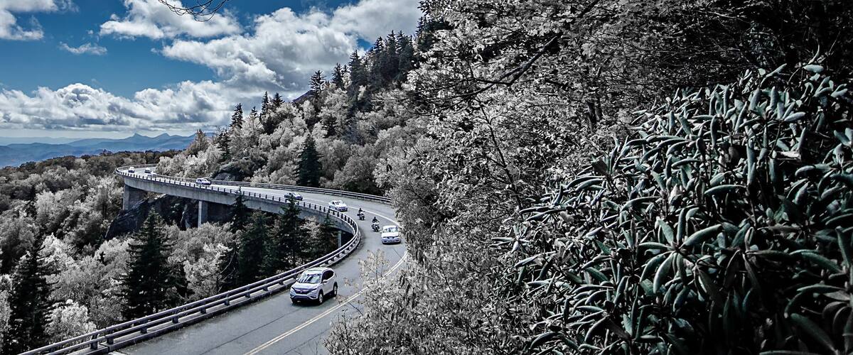 A winding mountain road with vehicles, surrounded by dense forest and distant mountains under a partly cloudy sky.