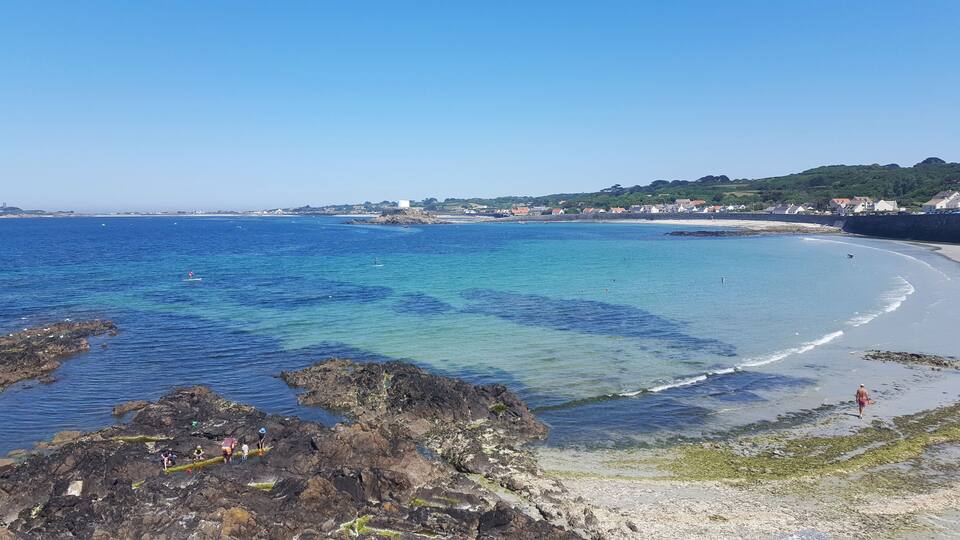 A coastal landscape with clear blue waters, rocky shores, and a sandy beach.