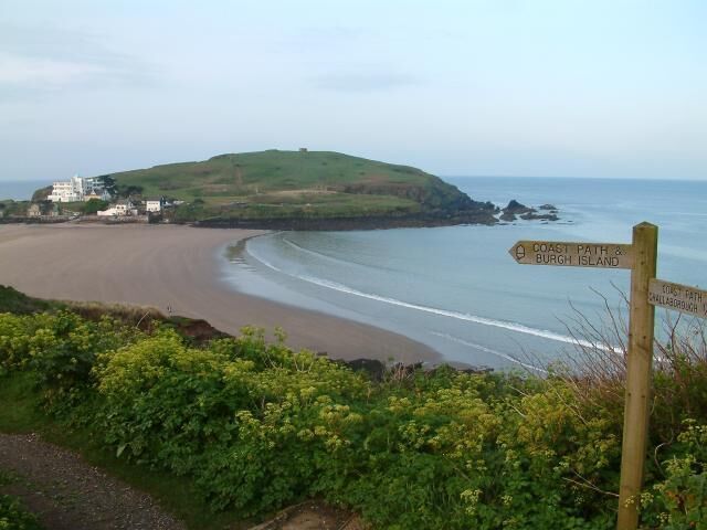 A coastal path sign, beach, and a hill with buildings in the background.