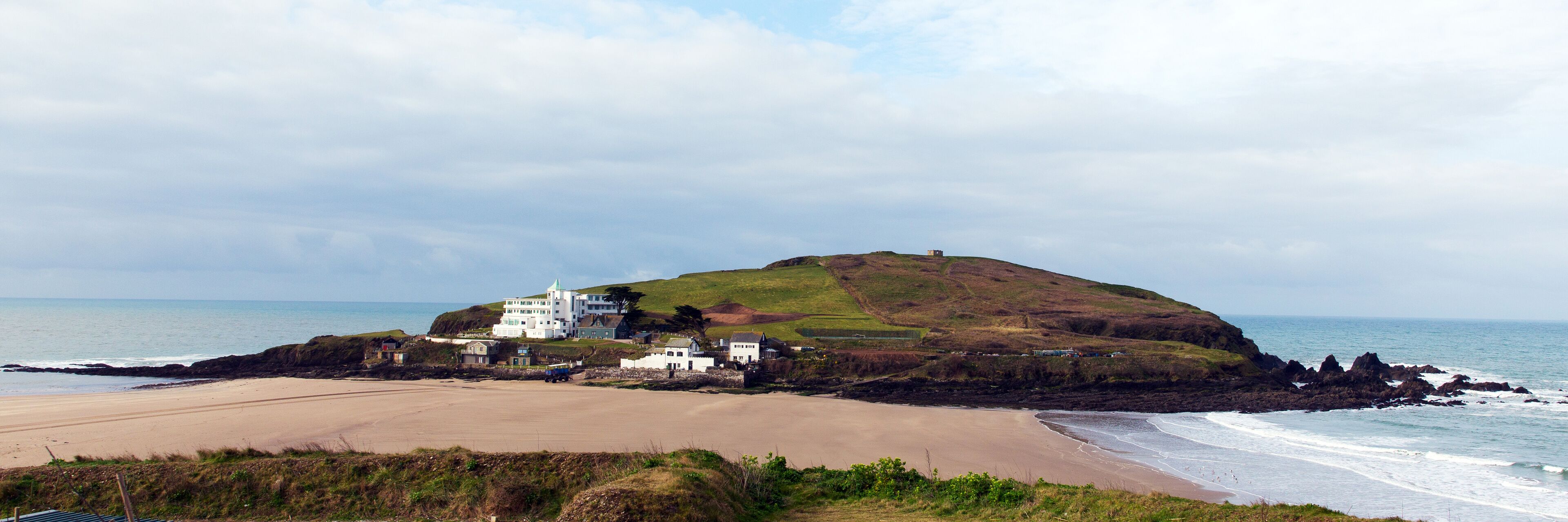 A coastal landscape with a hill, beach, and a white building complex.